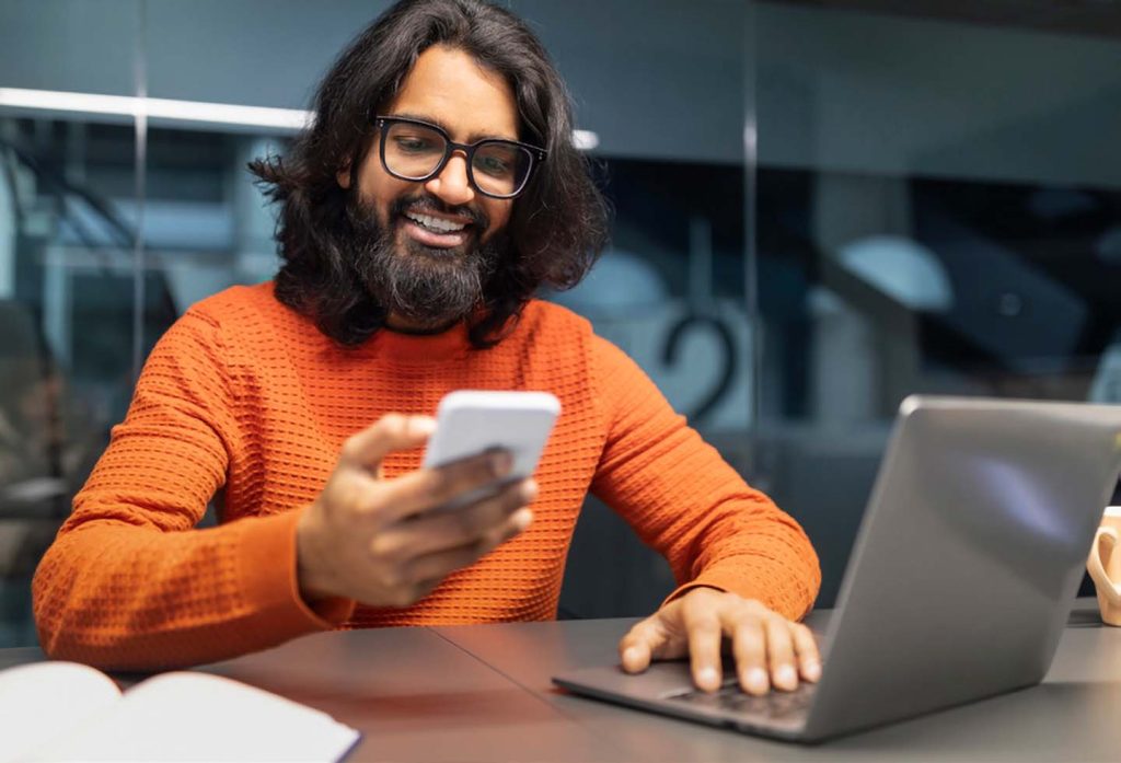 Cheerful Millennial Indian Man Using Smartphone and Laptop